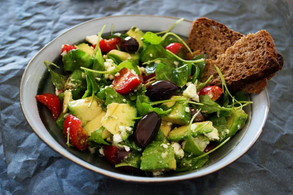 Gesunder Salat mit frischen Zutaten wie Avocado, Tomaten, Oliven, Feta und Rucola, serviert in einer Schüssel mit zwei Scheiben Vollkornbrot.